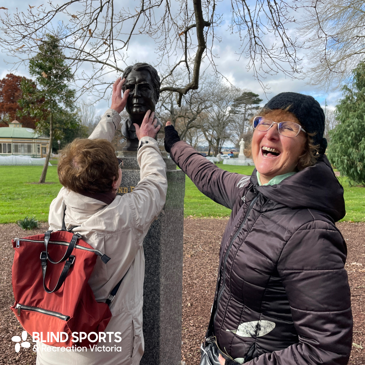 Carmel is standing with a BSRV participant who has both hands on the face of a bronze bust of a Prime Minister at the Ballarat Botanical Gardens. Carmel has turned to the camera and has a giant smile on her face. 